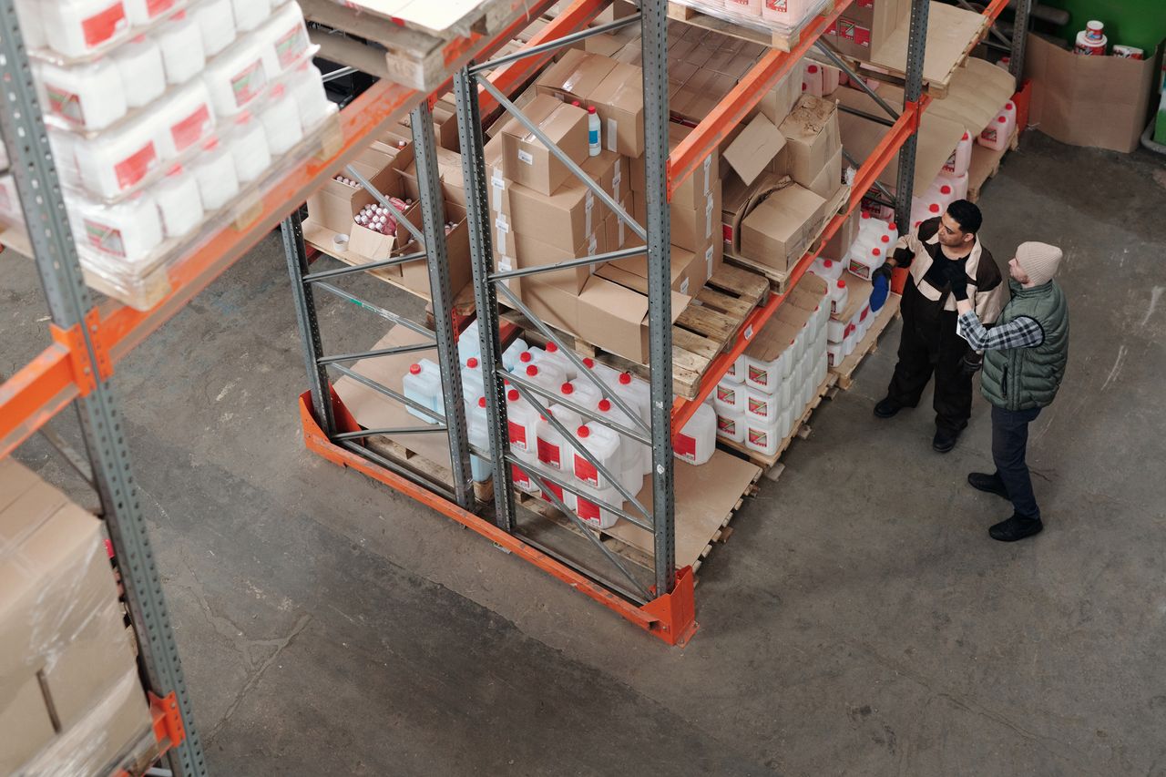 services-01 Two warehouse workers discussing logistics beside shelving with boxes and containers.