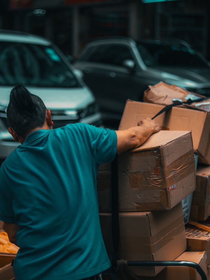 journey-01 Back view of unrecognizable young male worker in casual clothes loading stack of cardboard boxes on trolley while standing on street
