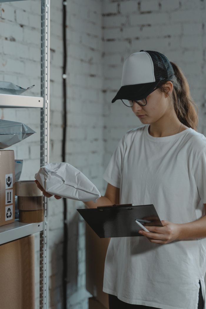 services-01 Young woman in white shirt and cap organizing packages in warehouse setting with clipboard.