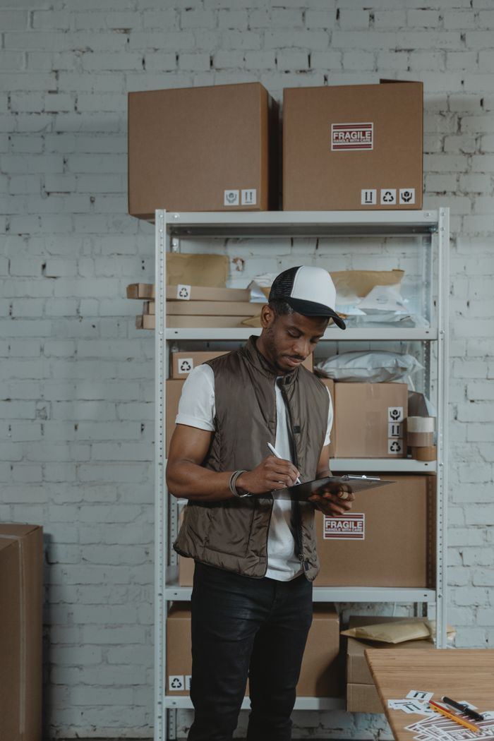 about-img Courier in a warehouse checking inventory on a tablet amidst stacked cardboard boxes.